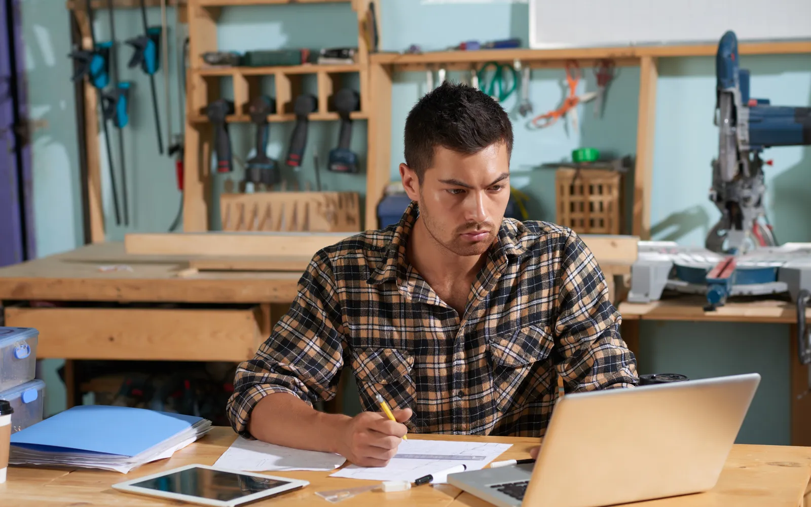 A joiner working at a bench with a laptop and drawings open in front of him