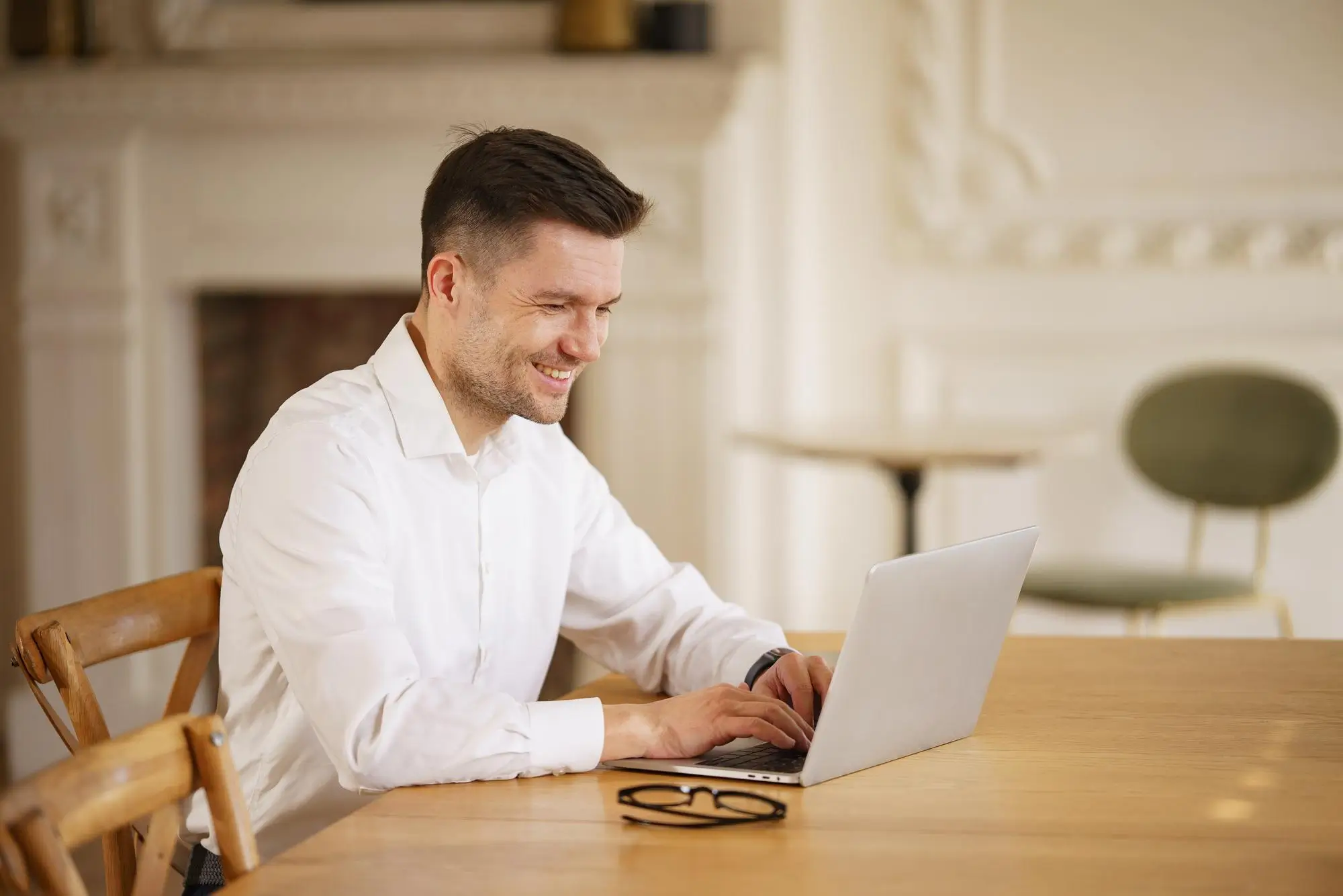A man in a white shirt smiling as he works at a laptop at a wooden table in a bright, airy room